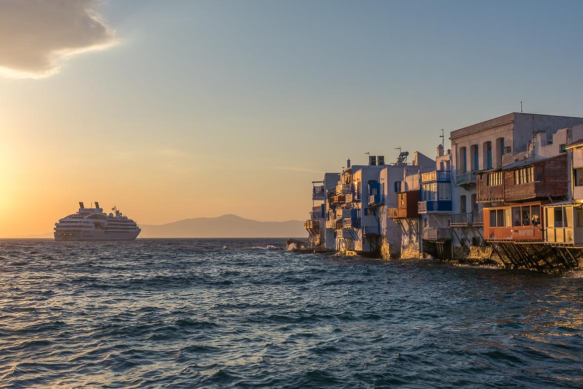 This image shows the row of beautiful buildings of Little Venice in Mykonos at sunset. The walls are painted gold and there is a cruise ship in the background. 