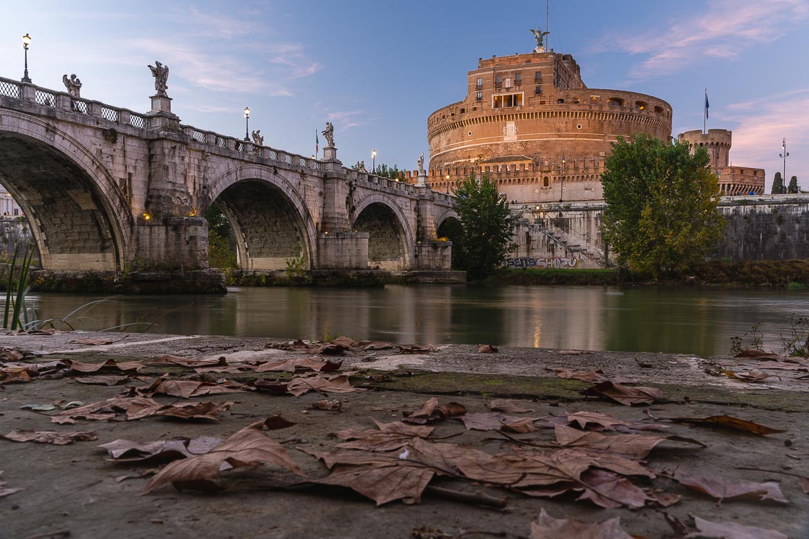 This image shows Ponte Sant'Angelo and the round Castel Sant'Angelo at sunset. In the foreground, the riverside path is covered with autumn leaves.