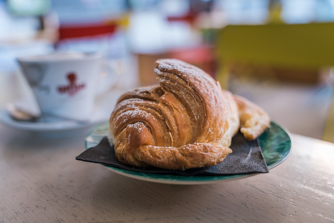 This is a close up of a cornetto, aka an Italian croissant. In the backround, blurred, a cup of cappuccino.