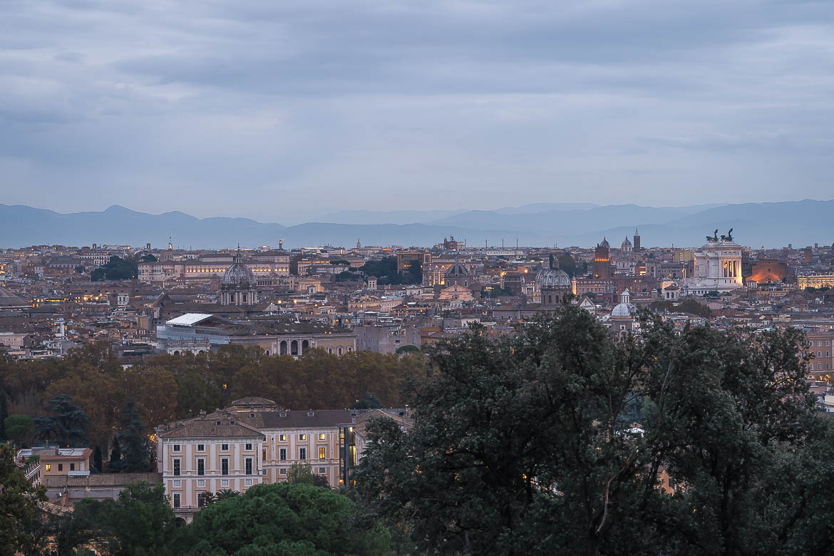 This image shows the view of Rome from the Belvedere of Gianicolo Hill at dusk. The skyline is stunning as the first city lights start to illuminate it.