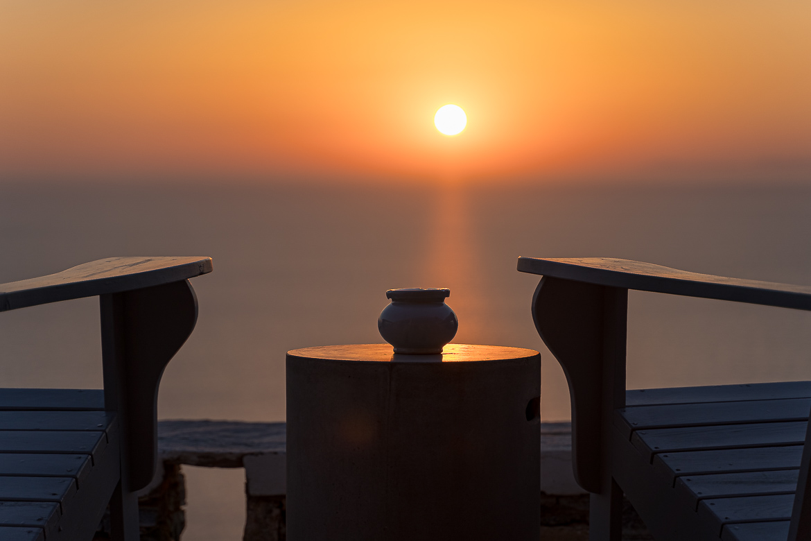 Τhis image shows the sun rising over the Aegean Sea. In the foreground, two wooden chairs and a table. If you're serious about travel photography, you need to be patient to catch the perfect shot. 