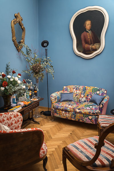 This image shows the cosy sitting area at Navona My Home. There's a colourful sofa, a red and white armchair and two vintage chairs. There are also a vintage painting and a mirror hanging on the walls, as well as a table with drinks, sweets, books and flowers.