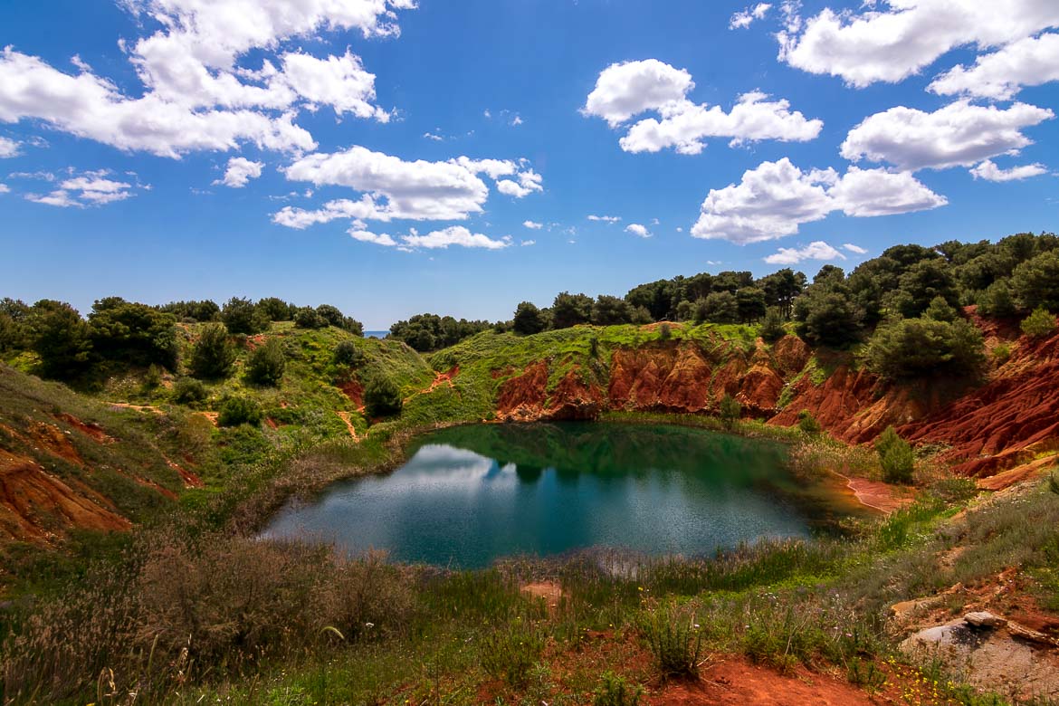 This is a photo of the bauxite lake. The lake's colour is green. It is surrounded by red rocks as well as trees. Above, a bright blue sky with only a few white clouds. 