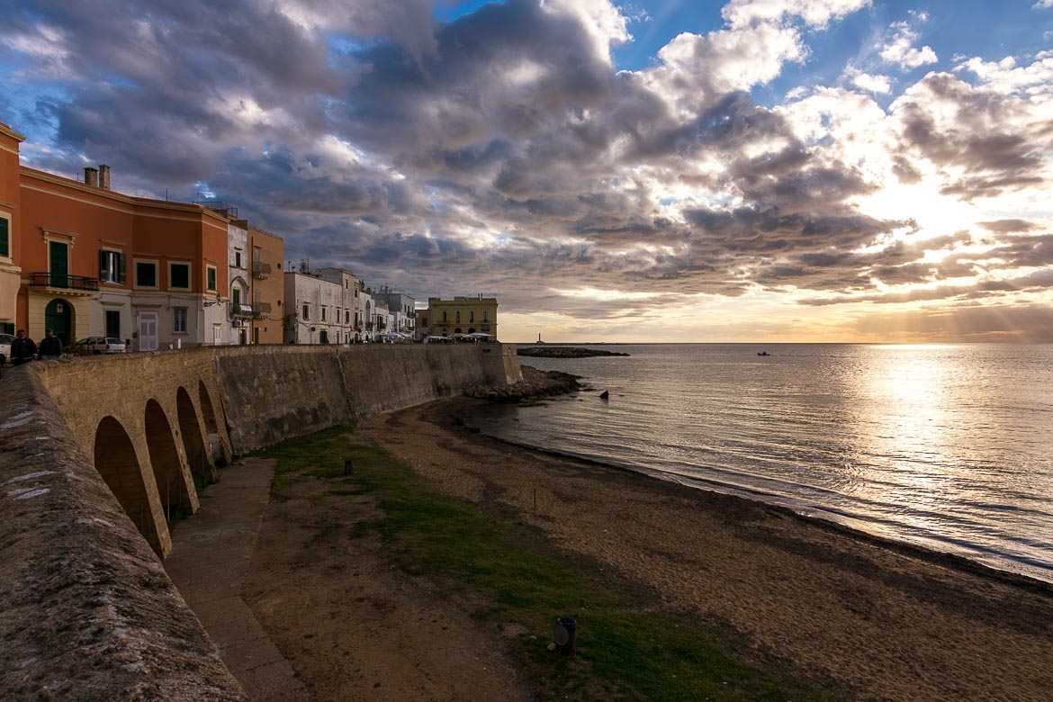 This photo shows the seafront promenade and the beach in Gallipoli at sunset. There is a cloudy dramatic sky as the sun dives into the Ionian Sea. 