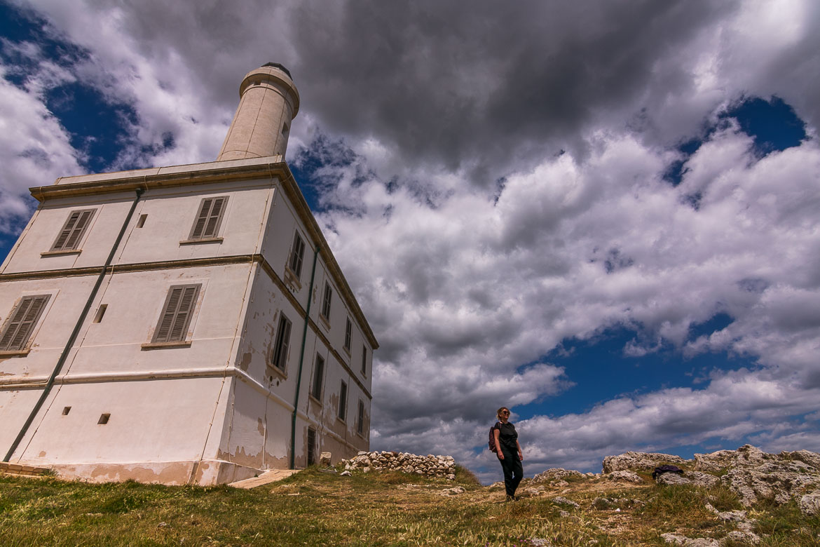 This photo shows the Punta Palascia Lighthouse. Maria is walking in front of it with a wide smile on her face. 