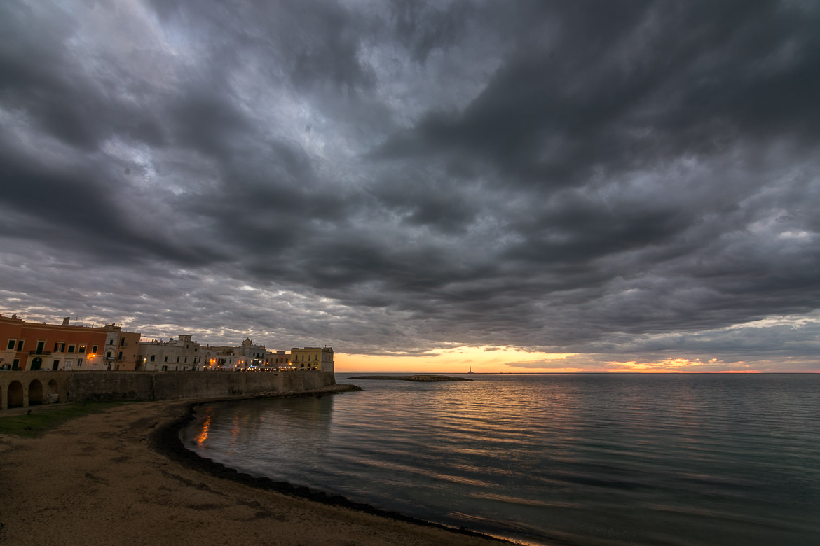 This is a panoramic shot of Gallipoli beach at sunset. The sky is stunning with grey clouds over the orange setting sun. To the left, the old palazzi of Gallipoli dimly lit look beautiful. 