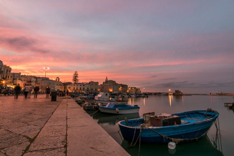 This image was shot in Trani port during sunset. The sky is a magnificent collection of pinks, yellows and blues, all reflecting on the mirror-like sea. There are small blue fishing boats in the sea and people walking along the promenade. The old-fashioned palazzi are dimly lit. We chose this photo to be the featured image for our article: Best beach towns in Puglia Italy for a laid-back vacation.