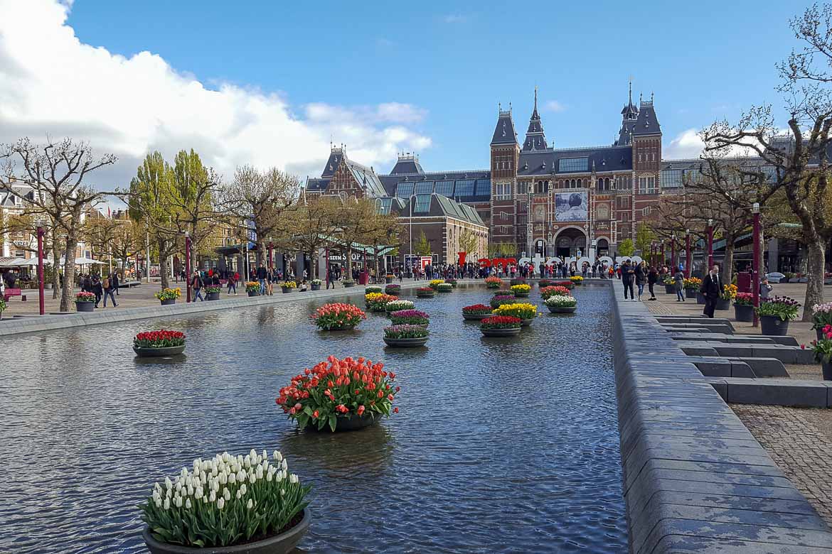 This image shows the Rijksmuseum in Amsterdam. It is a gorgeous ornate red building with an I love Amsterdam sign in front of it as well as a beautiful canal decorated with flowers.