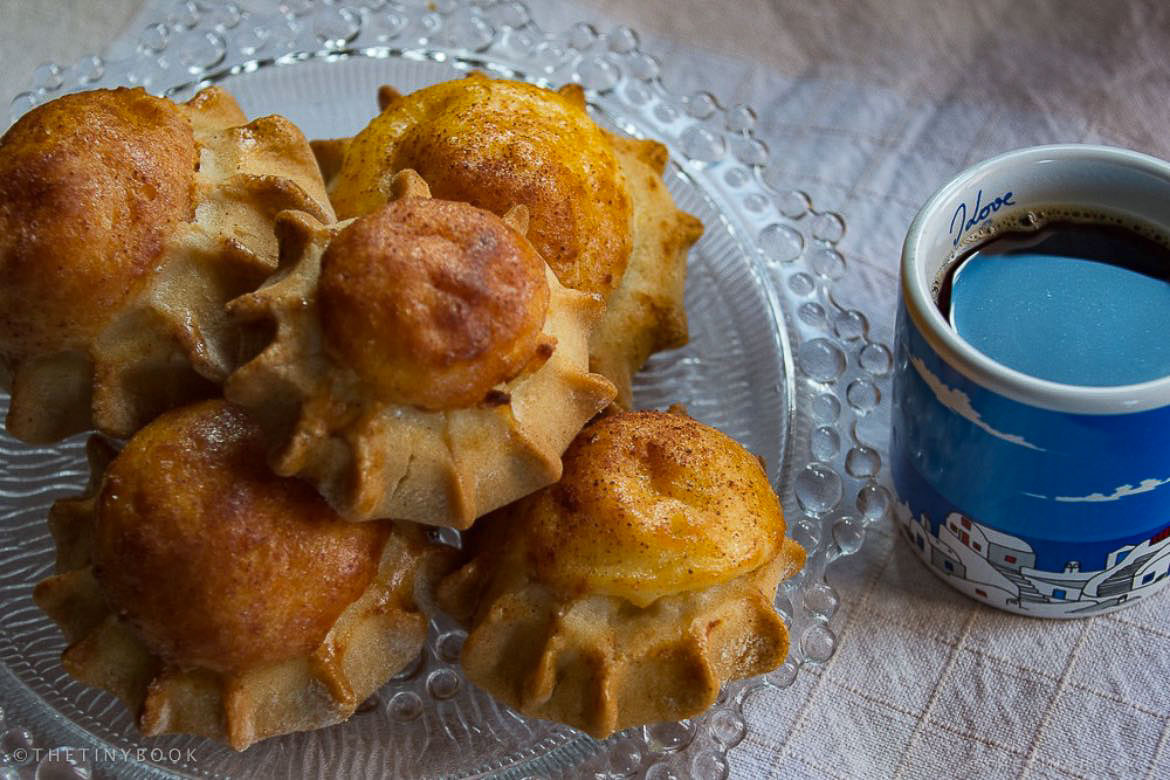 This is a close up of a traditional Cretan Easter treat which is called Lychnarakia. It's a kind of pastry filled with local sweet cheese and sprinkled with cinnamon. Next to the plate of Lychnarakia there is a mug of coffee.