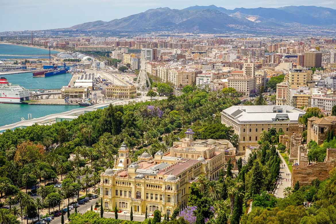 This is a panoramic shot of Malaga. In the foreground, there is a beautiful boulevard lined with palm trees and other trees as well as gorgeous historical buildings. In the background, the modern city as well as part of the port are visible.