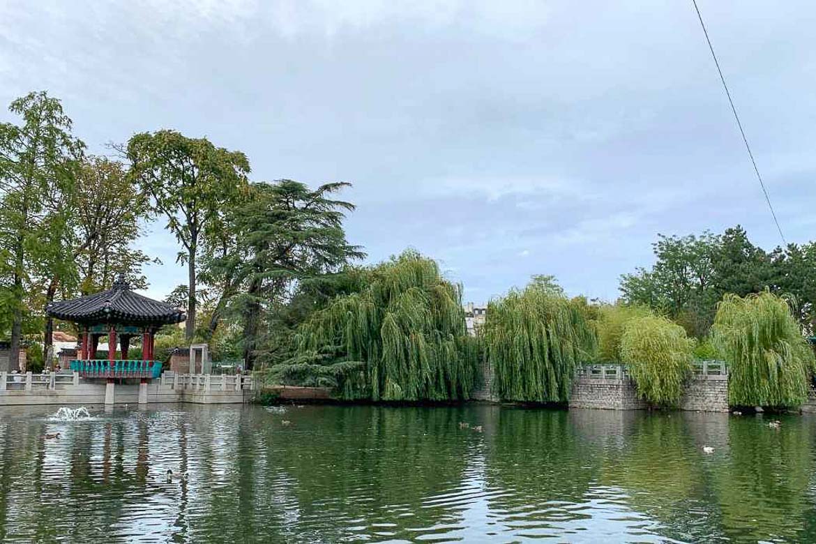 This image shows a small lake on which ducks float in Jardin d' Acclimatation in Paris. The lake is surrounded by trees.
