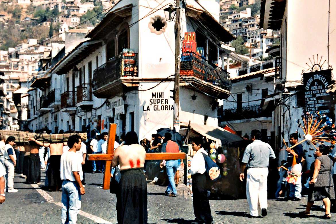 This image shows part of the procession of penitents in Taxco, Mexico. There is one man with his bloodshed back turned to the camera who is carrying a huge wooden cross. There are many spectators at the side of the street.