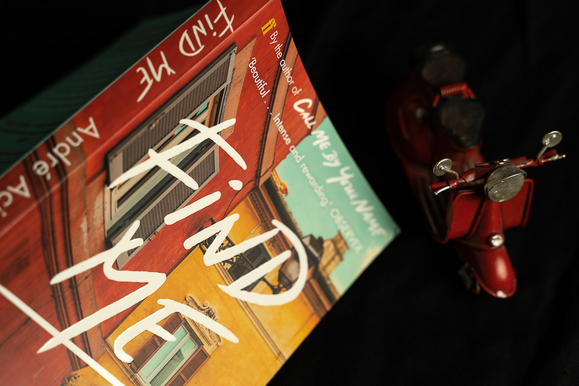 This image shows a paperback edition of Find me by Andre Aciman next to a red toy vespa scooter.