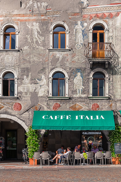 This image shows a beautiful facade in Trento. There's also a traditional Italian cafe where people are sitting on a sunny day.