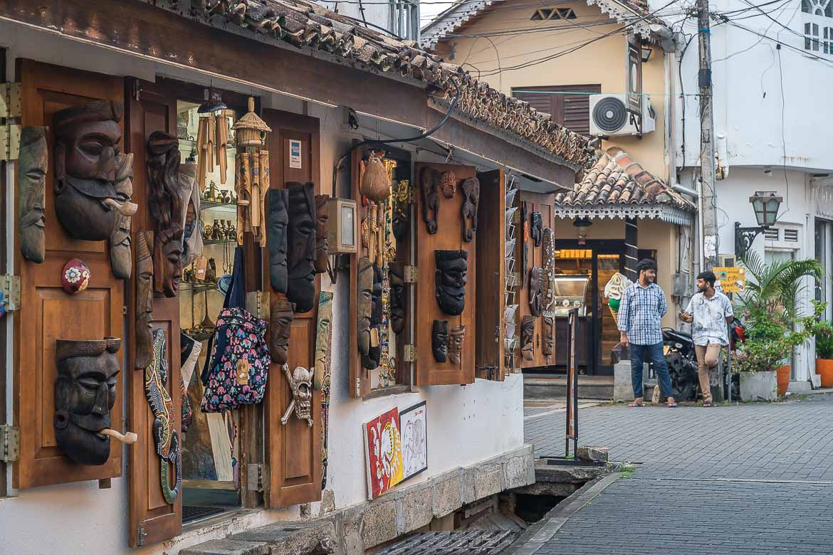 A souvenir shop with wooden masks and crafts hanging on the shutters. Two locals are chatting on the street across the shop.