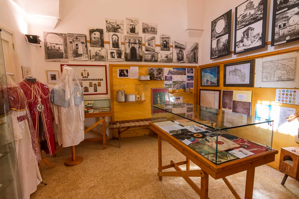 This photo shows various displays inside the folk museum in Calimera. There are three traditional costumes as well as old LPs, maps and black and white photos. 