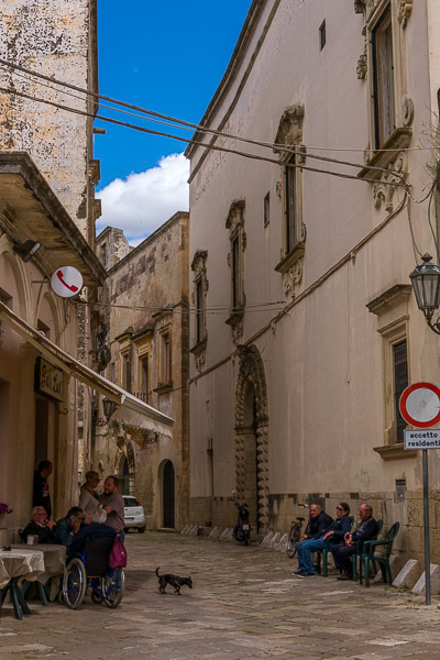 This image shows a quaint street in Corigliano d'Otranto. People are sitting on chairs on both sides of the street chatting. 