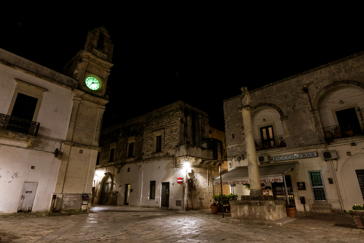 This is a night shot of Corigliano d'Otranto. There is a clock tower and a closed Italian bar. 