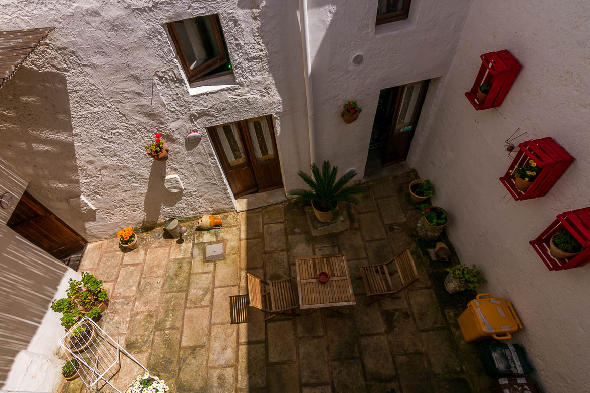 This photo was shot from above and shows the casa a corte. This architecture style is typical of this area in Italy. We can see two doors opening to the same courtyard. There is a table, chairs and flower pots there. 