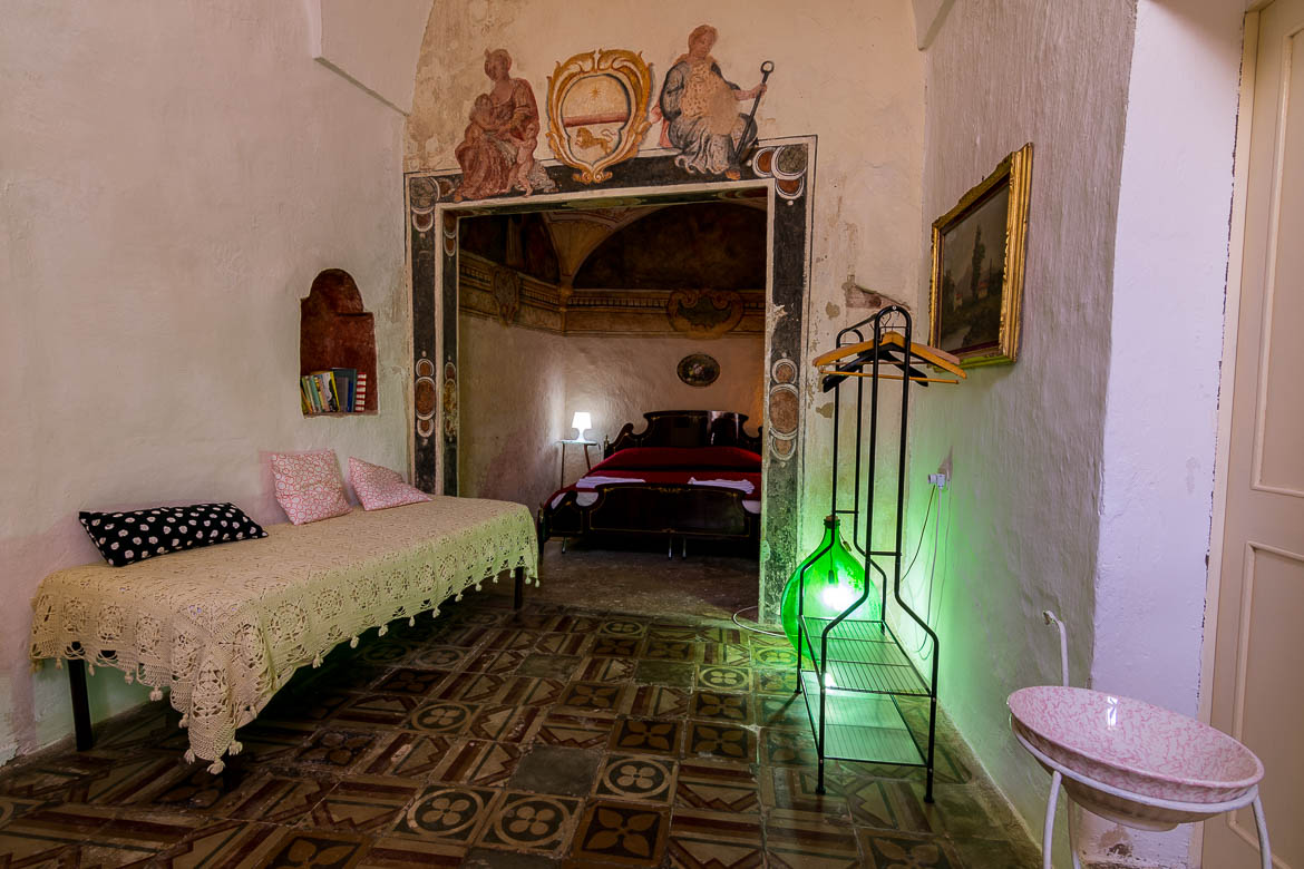 This room shows the interior of a room at Corte Candelora B&B. The furniture is old-fashioned and there is a green lamp. The floor has the same tiles it used to have since its construction. On the walls, there are painted frescoes with religious themes. 
