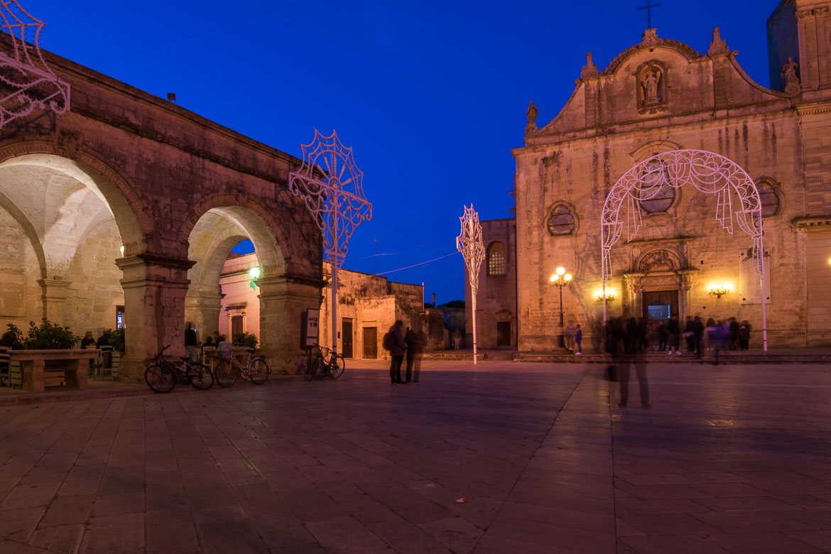 This is a photo of Piazza san Giorgio in the evening. The church and the market building are lit and the sky is light blue. 