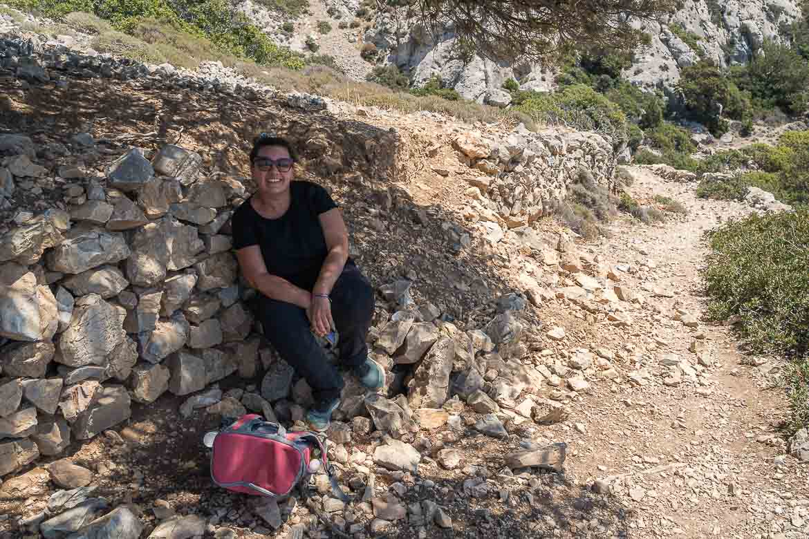 Maria is resting on a stone under a tree shade on the way back from the Cave of Saint John.