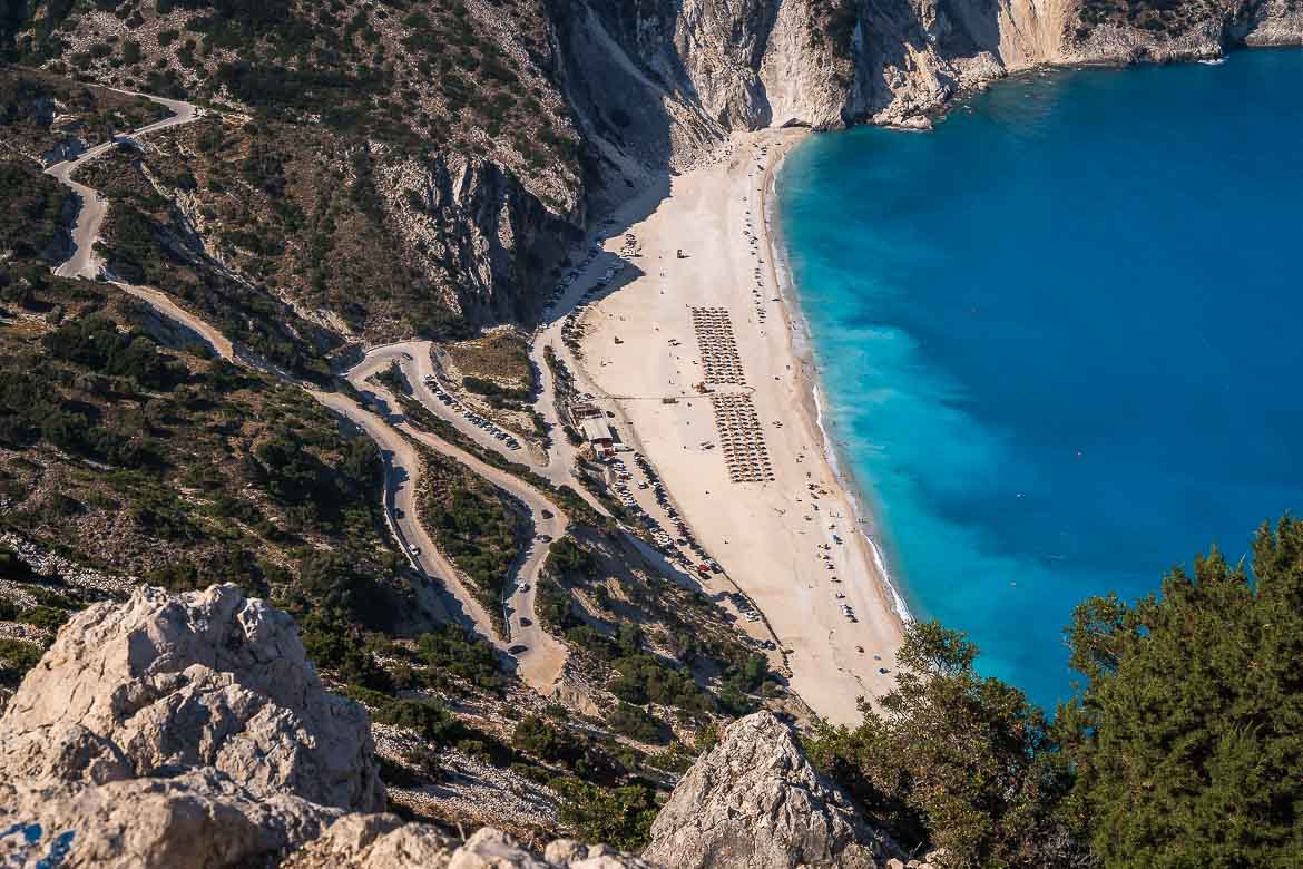 Panoramic view of Myrtos beach in the morning when the water is deep blue. At the end of the winding road that leads to the beach, there are a lot of cars parked.