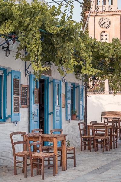 This is the facade of one of the most beautiful cafes in Lefkes, to Kafeneio tis Marigos. The doors and shutters are painted light blue and there are brown tables and chairs outside, right underneath a lush vine.