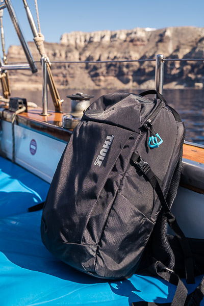 This image shows a Thule backpack on board a sailing boat in Santorini. In the background, we can see the island's iconic cliffs. A minimalist travel photography gear list is never complete without a good minimalist travel backpack.