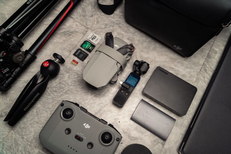 This image shows some items from Katerina's minimalist travel photography gear: a travel tripod, a pocket-sized tripod, a small drone, the drone joystick, two hard drives, four SD cards, the DJi Osmo Pocket, an ultrabook and a compact shoulder bag.