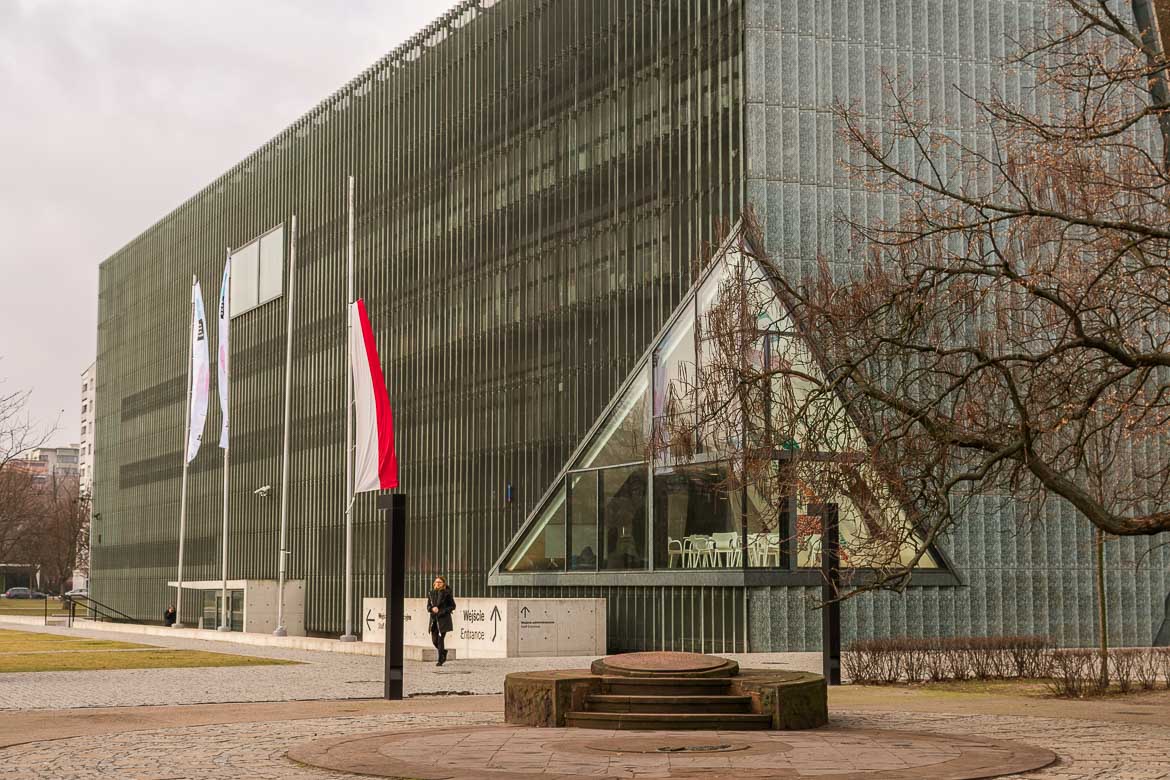 The austere facade of POLIN Museum. The building is made of glass and concrete. Outside, there's a flag of Poland. There's also a woman dressed in black walking around.