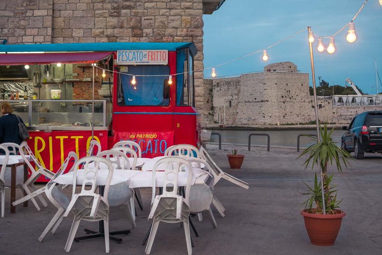 This photo shows a food truck on Trani promenade selling fried fish. The food truck is red and there is a line of lights hanging above. There are white tables and chairs. This photo is indicative of the simplicity of food in Puglia and this is why we chose it as the featured image for our Puglia food guide.