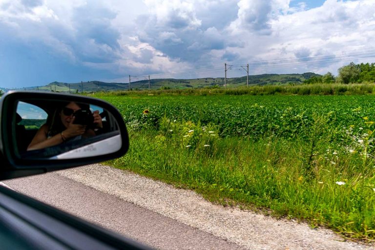 This image shows the green Romanian countryside as we drive along a road. There is the reflection of Katerina shooting the photo on the car mirror. This is the featured image on our Romania road trip article.