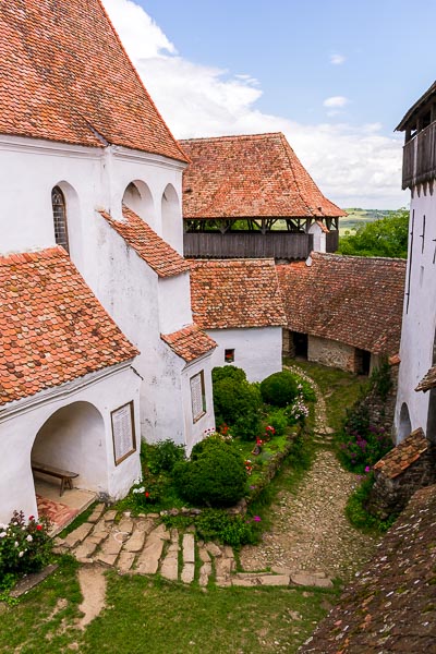 This image shows the interior of the fortified church in Viscri. It is a complex of buildings which look like one. The church is surrounded by green plants and pretty flowers. The walls are white and there are red tiled rooftops.
