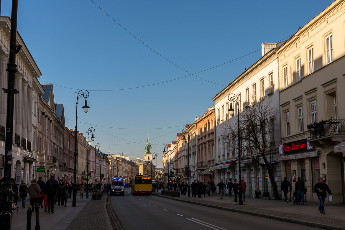 Nowy Świat Street is one of the streets that comprise the Royal Route in Warsaw Poland. It's lined with nice buildings. There's traffic on the road and people walking on the sidewalks.