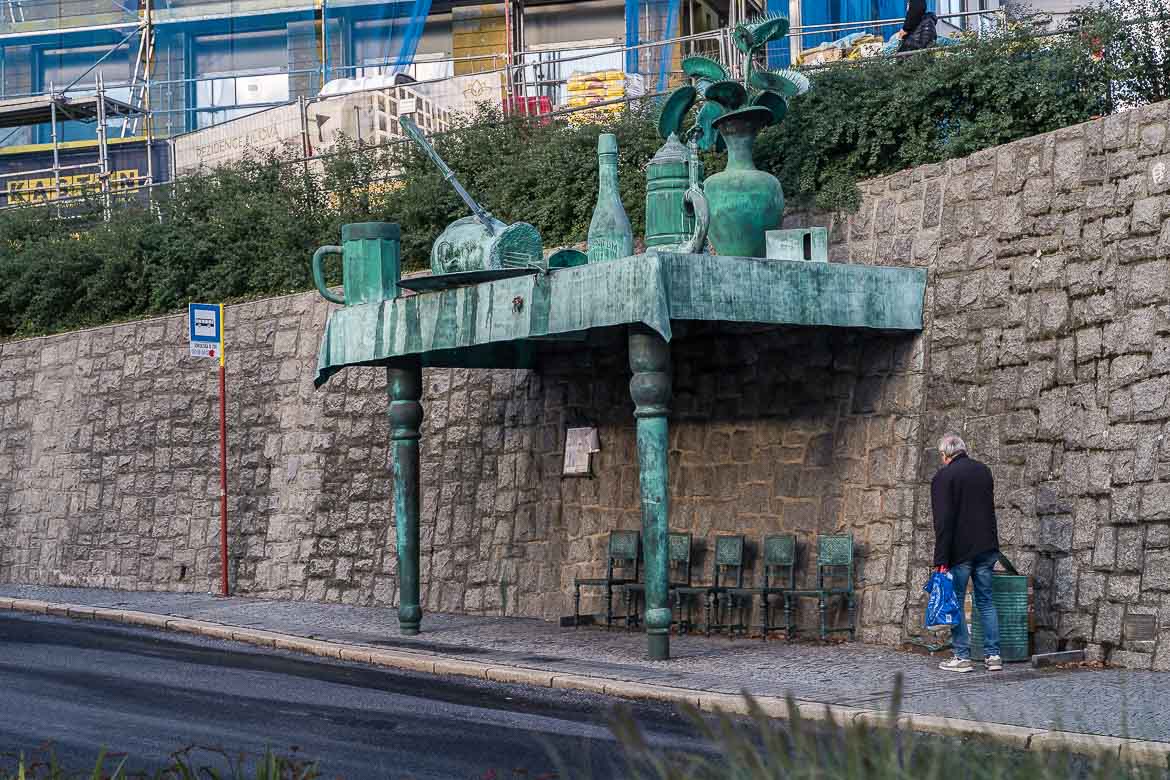 This image shows a bronze bus stop which is called The Feast of Giants. There are five bronze chairs for people to sit while waiting the bus. On the roof of the bus stop, there are bronze objects like a beer, a bottle, a plant etc. A man is walking towards the bus stop.