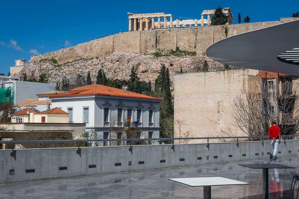 View of the Parthenon from the Acropolis Museum terrace. A man with a red jumper walks around the terrace.