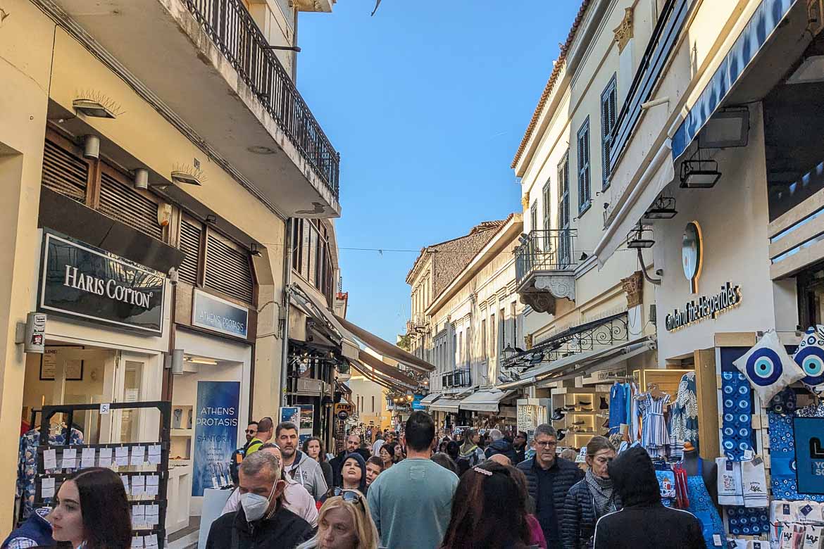 Crowds on Adrianou Street in Plaka on a sunny day. The street is lined with commercial shops.