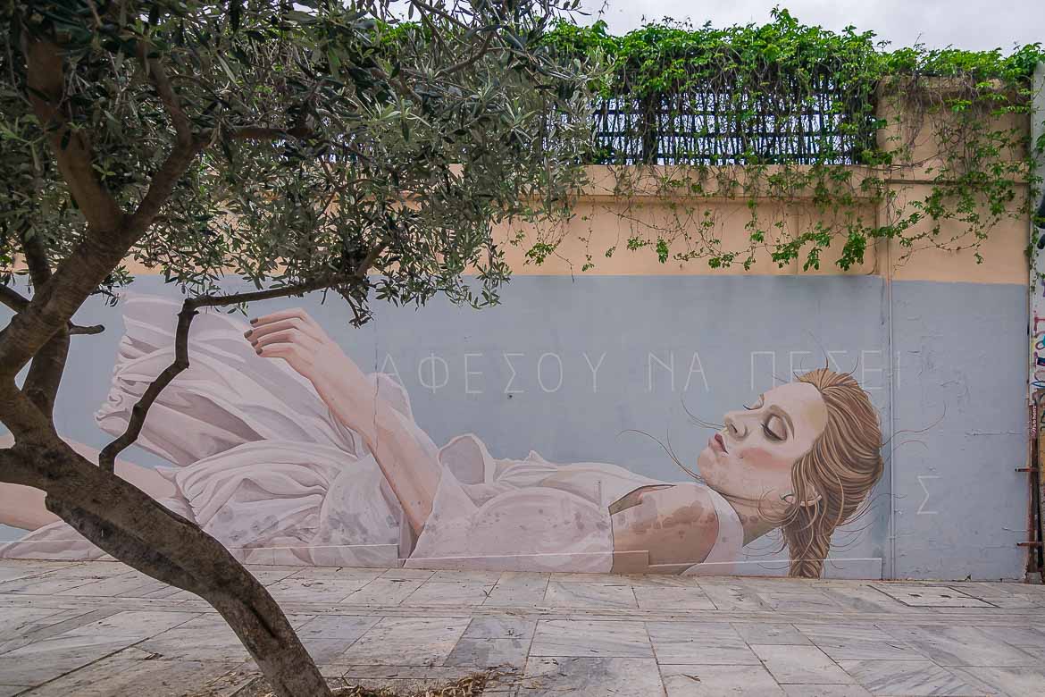 A street artwork on a wall showing a blonde woman wearing a white dress. On the wall, it's written in Greek: let yourself fall.