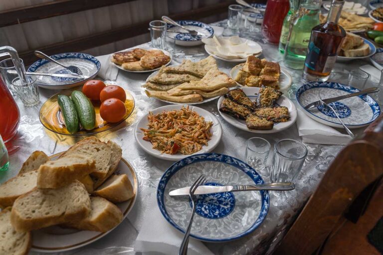 A table laid for lunch. There are white and blue plates, bread, tomatoes, cucumbers, Khachapuri, water and wine.