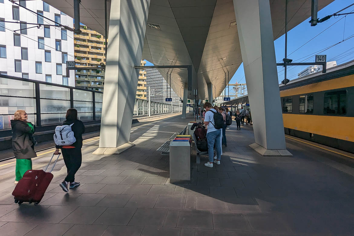 Several passengers with luggage on the main train station's platform. A yellow train has arrived at the railway on the platform's right side.