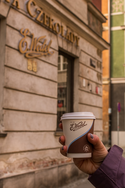 This image is a close-up of Maria's hand holding a paper cup of hot chocolate outside E. Wedel café.