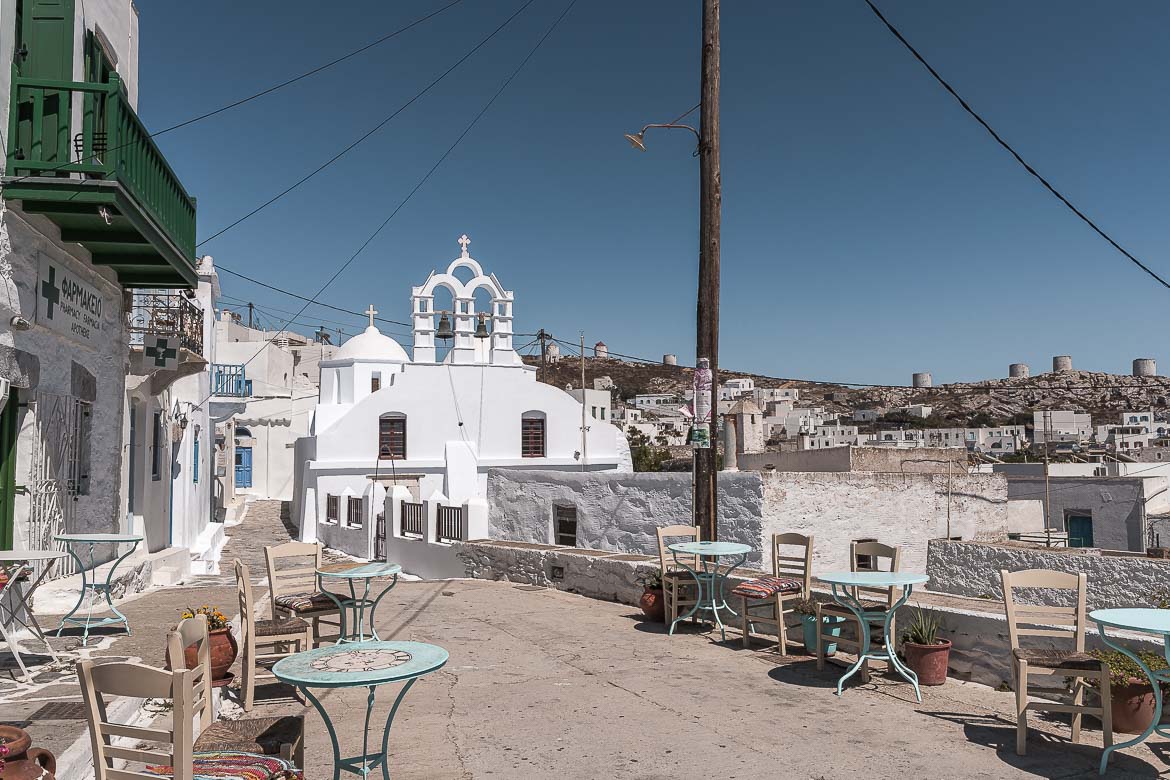 This image shows the entrance to Chora, the old town of Amorgos. There is a church dominating the photo and traditional tables and chairs lining the street.