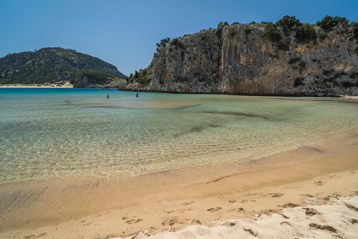 View of Voidokilia beach from the sandy shore. The turqoise waters are crystal clear and the beach is surrounded by dramatic rocky cliffs.