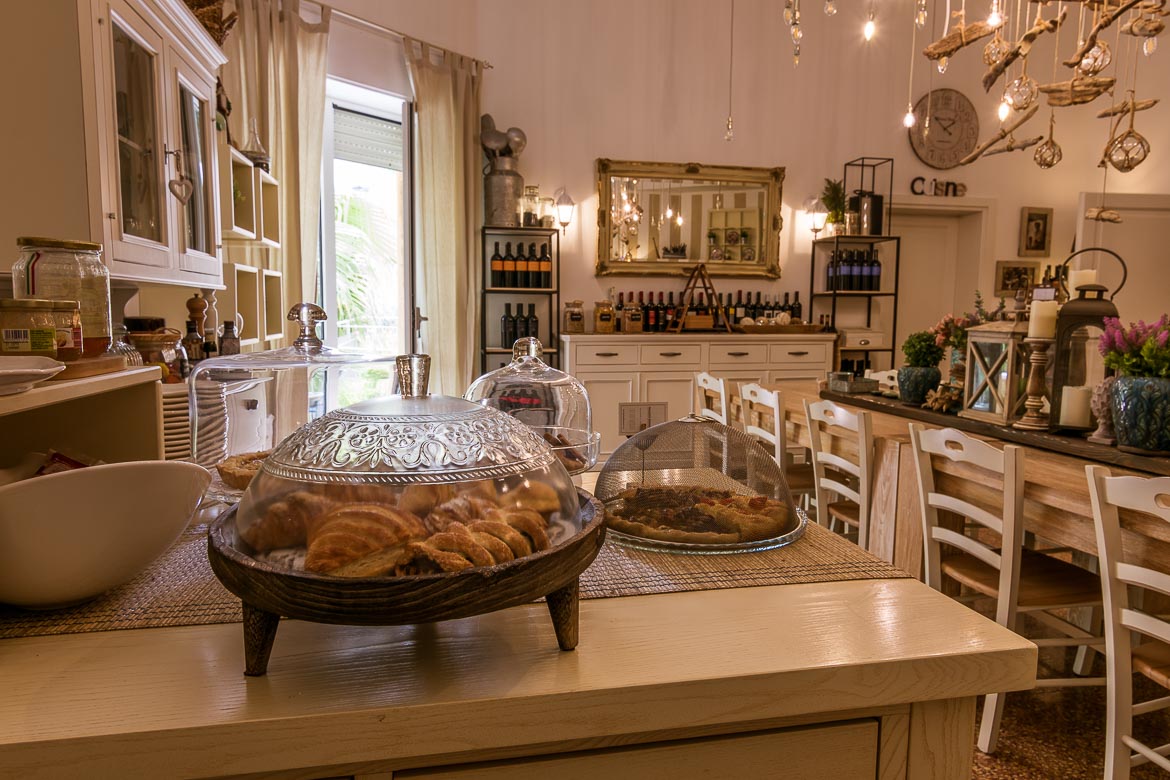This photo shows the kitchen at Palazzo Bignami. In the foreground, platters with sweet and savoury treats. In the background, the cosy dining table with chairs and the wine collection. 