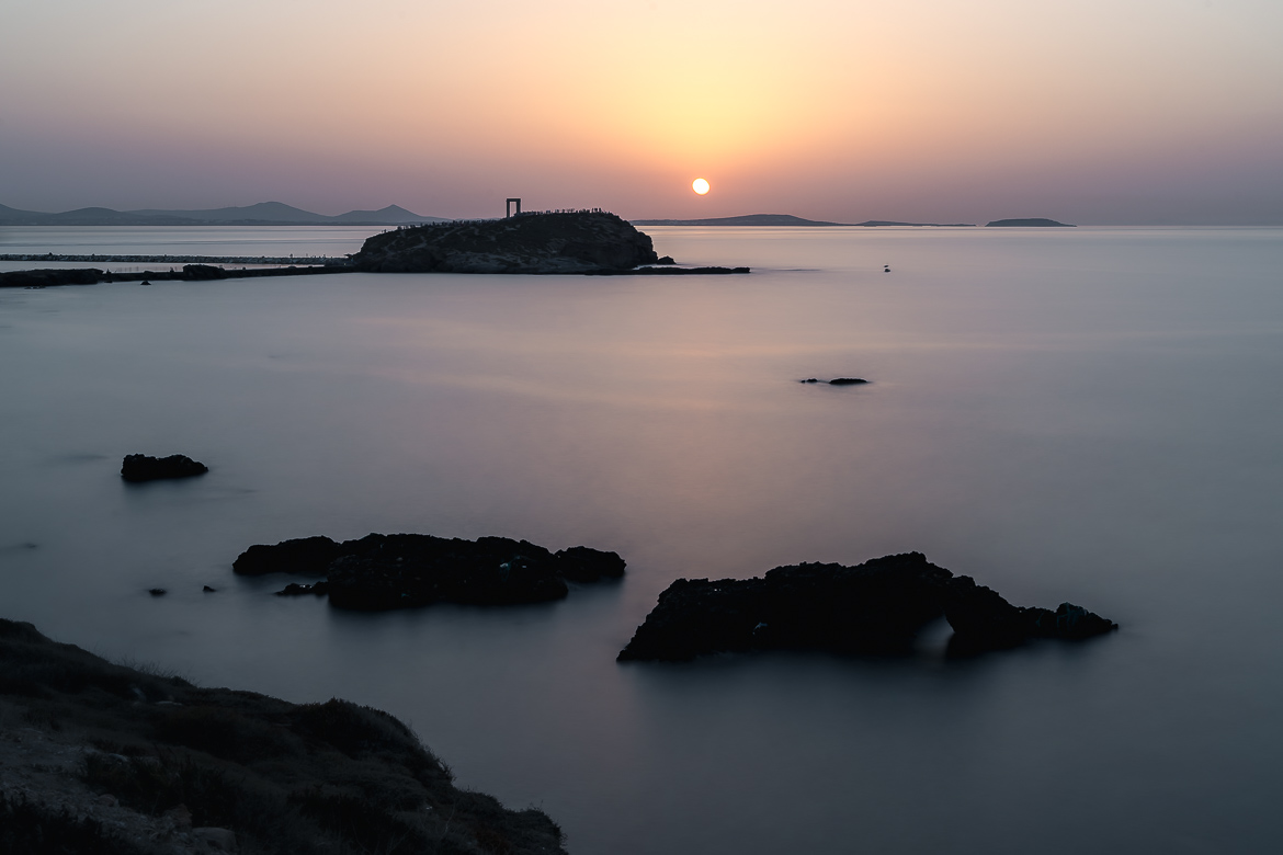 This image shows Portara during sunset. The photo is taken from Grotta and the sea is smooth and calm.