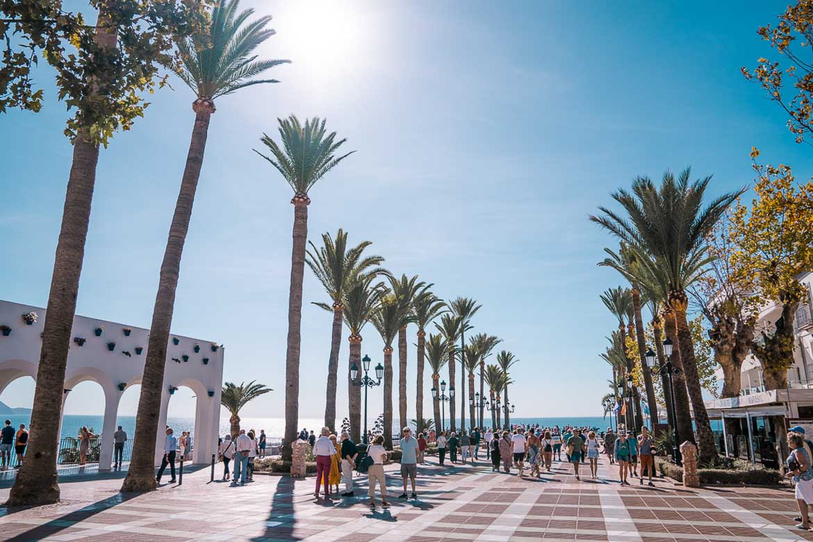 This image shows people walking along a palm tree-lined wide boulevard at the Balcon de Europa Nerja. 