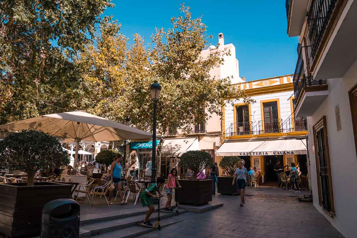 This image shows a quaint square. There are people sitting at tables or walking. 