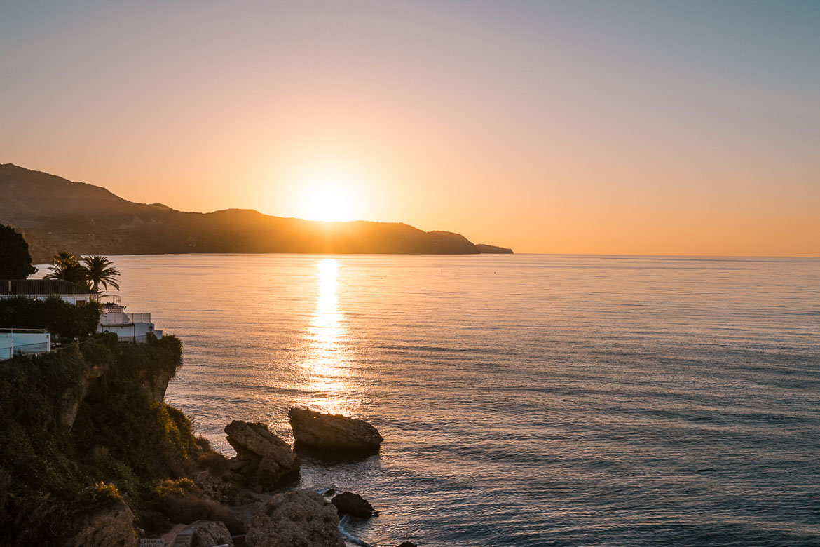 This images shows the sun rising behind a mountain. In the foreground, the sea and some rocks. 