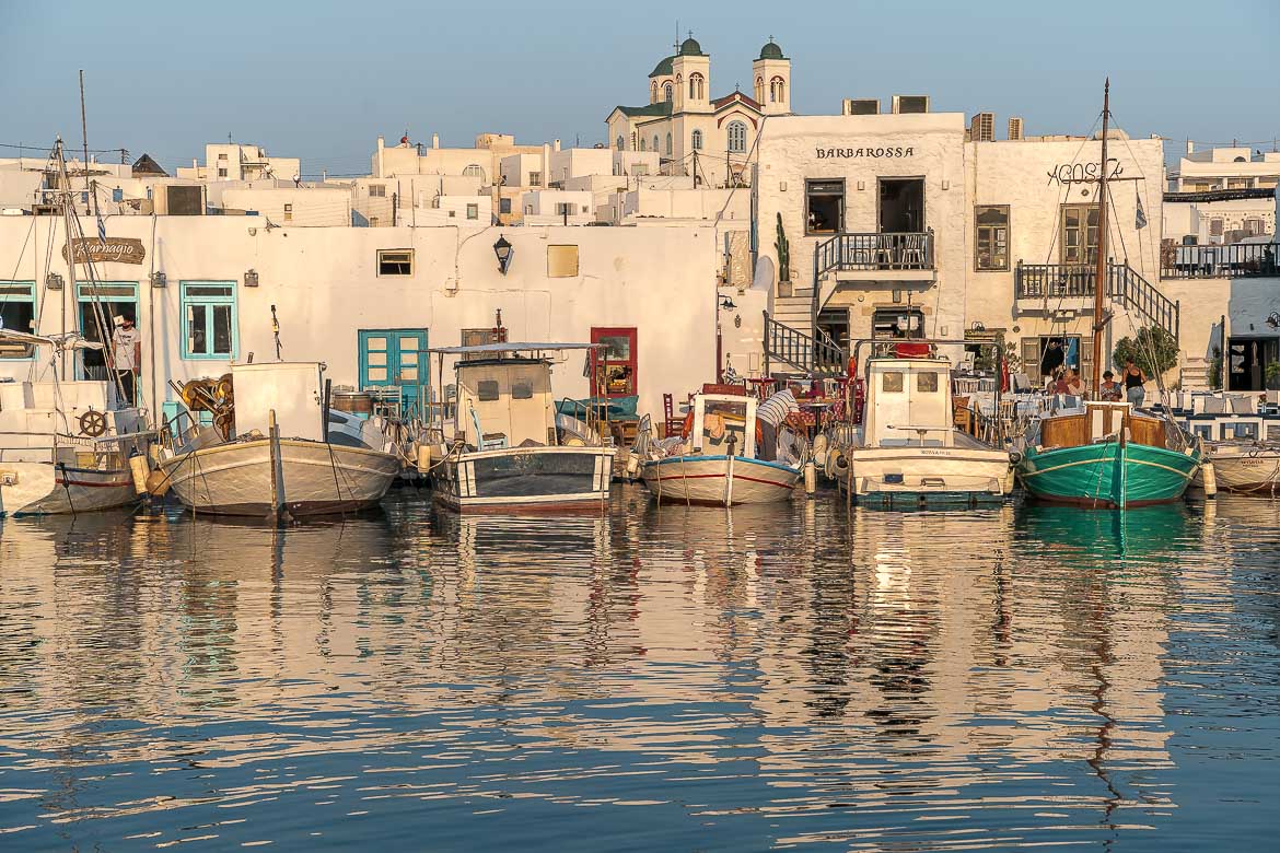 This is a panoramic shot of the Old Port in Naoussa at sunset. There are many traditional boats which reflect on the calm sea water. In the background, the whitewashed buildings of Naoussa. 